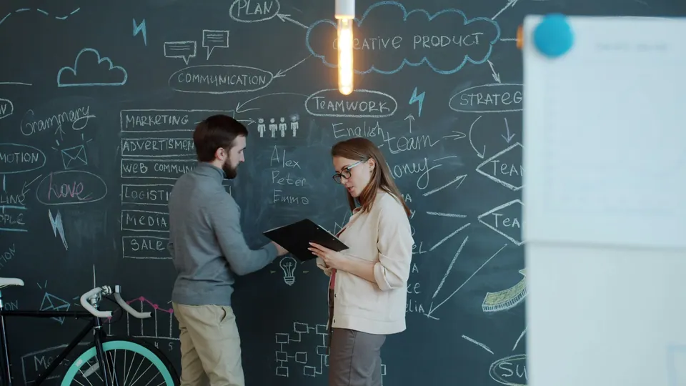 Two colleagues reviewing a document in front of a chalkboard with strategy and teamwork notes